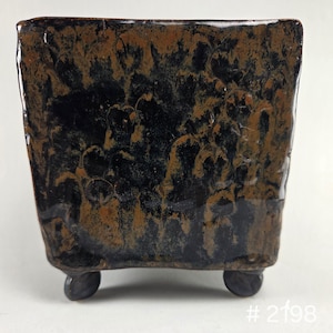 May include: A square ceramic planter with a glossy, mottled glaze in shades of deep black and earthy brown. The vessel features a textured, uneven surface and is supported by four small, rounded feet at the base. The text # 2198 is visible.