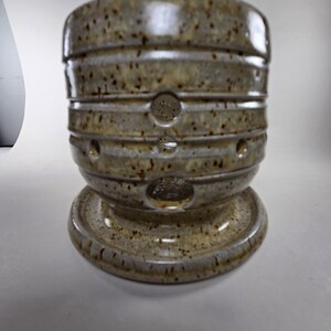 May include: A handcrafted ceramic vessel with a speckled, earthy glaze. The piece features a cylindrical body with horizontal ridges and circular openings. It sits on a rounded base, showcasing a unique, organic design. The color palette includes shades of tan, brown, and gray.