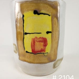 May include: A ceramic mug with a white base and a yellow and brown abstract design. The design features a yellow square with black lines and a red square. The number "# 2104" is printed at the bottom.