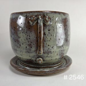 May include: A ceramic planter with a saucer, featuring a speckled sage green and brown glaze. The exterior has a raised T-shaped relief with two circular accents at the top and one at the bottom. The text # 2546 is visible in the bottom right corner.