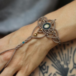 Könnte beinhalten: Handgefertigtes Makramee-Armband in Beige, mit einem ovalen Stein in der Mitte, umrandet von Dunkelblau. Das Armband hat ein Blumenmuster, kleine goldene Perlen und einen Fingerriemen. Ein einzigartiges Schmuckstück.