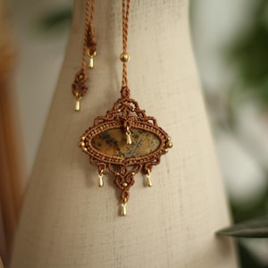 Può includere: Una collana macramè fatta a mano con una pietra ovale centrale con motivi marroni e beige. Il ciondolo è incorniciato da intricati nodi macramè in un caldo colore marrone, accentuato da perline dorate e dettagli pendenti.