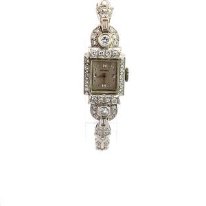 Op de afbeelding: Een vintage polshorloge met een vierkante wijzerplaat en een band bezet met diamanten. De wijzerplaat is wit met zwarte Romeinse cijfers als uurmarkeringen en het woord "LATEMINE" staat in het zwart gedrukt.