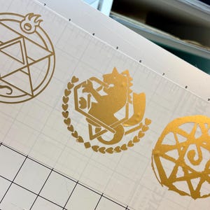 May include: Gold-colored vinyl decals featuring three distinct designs. One is a circular emblem with a lizard and flame, another depicts a wolf-like creature within a heart-shaped wreath, and the last is a spiral design.