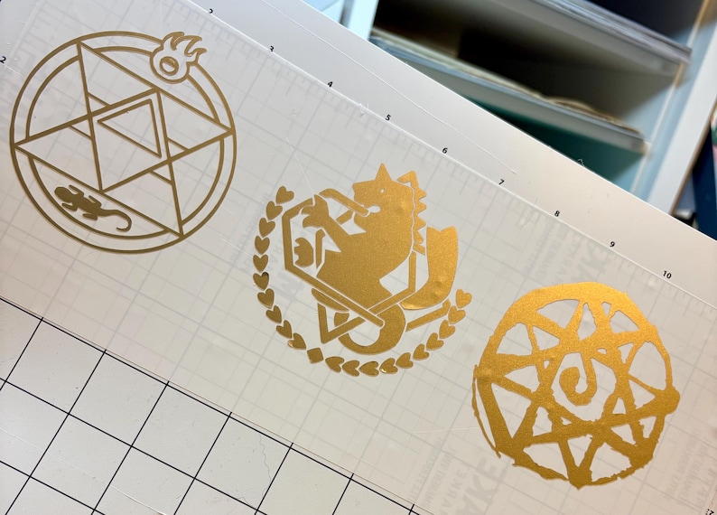 May include: Gold-colored vinyl decals featuring three distinct designs. One is a circular emblem with a lizard and flame, another depicts a wolf-like creature within a heart-shaped wreath, and the last is a spiral design.