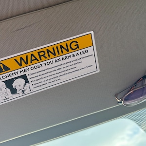 A car visor with a warning sticker that reads "WARNING ALCHEMY MAY COST YOU AN ARM & A LEG." The sticker has a yellow and black border and a picture of the Elric brothers performing human tranmutation. A pair of purple-tinted glasses is clipped to the visor.