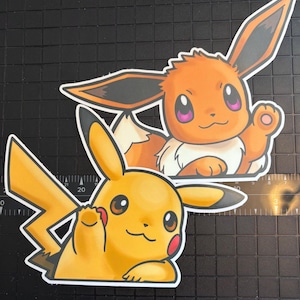 May include: Two die-cut stickers featuring popular Pokémon characters. One sticker depicts Pikachu, a yellow creature with red cheeks and a lightning bolt tail. The other sticker shows Eevee, an orange creature with large ears and a fluffy tail.