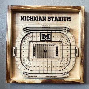 Puede incluir: Una pieza de arte de pared de madera con un diseño detallado de corte por láser del Michigan Stadium, también conocido como "The Big House". El estadio se representa con el icónico logotipo "M" y secciones de asientos numeradas. La pieza está enmarcada en un marco de madera rústico.