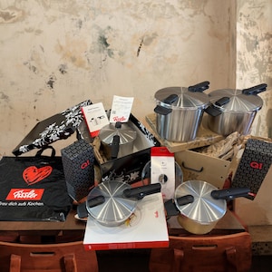 May include: A collection of stainless steel Fissler pressure cookers and accessories. The image shows several cookers with black handles and lids, along with packaging and a black shopping bag featuring a red heart logo. The cookers are on a wooden table.