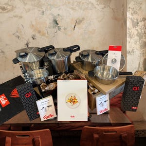 May include: An assortment of stainless steel Fissler pressure cookers and accessories are displayed on a wooden table. The set includes multiple pots, a steamer insert, and packaging with the Fissler logo and product information.