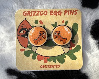 May include: Two round Grizzco Egg Pins with orange and black designs. Each pin features a cartoon egg with a jagged, orange creature inside. The pins are on a tan card with the text "GRIZZCO EGG PINS" and "OBICREATES".