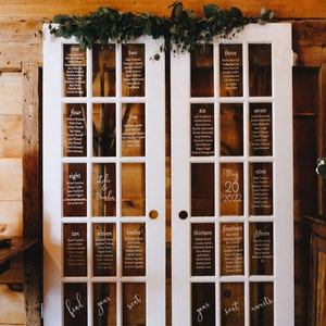 Peut inclure: Deux portes doubles blanches avec des vitres, décorées de verdure, utilisées comme plan de table de mariage. Les vitres sont étiquetées avec les numéros un à quinze, et chaque vitre liste les noms des invités. Les portes sont décorées avec le texte "find your seat" et "your seat awaits".