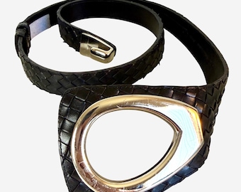 May include: A black woven leather belt featuring a large, asymmetrical, polished silver-tone metal buckle. The belt strap has a consistent braided texture throughout, and the buckle has a smooth, reflective surface with a teardrop-shaped cutout.