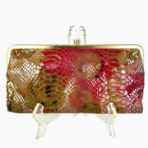 May include: A rectangular clutch bag featuring a textured snakeskin-patterned surface in shades of red, gold, and brown. The bag is finished with a metallic gold-toned frame and a small, centered cylindrical clasp at the top edge.