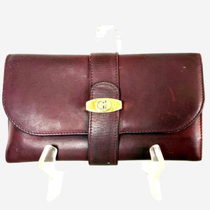 May include: A rectangular dark burgundy leather wallet featuring a central vertical strap secured by a gold-toned metal clasp. The clasp displays an embossed logo design. The wallet has a smooth finish with visible natural grain and stitching.