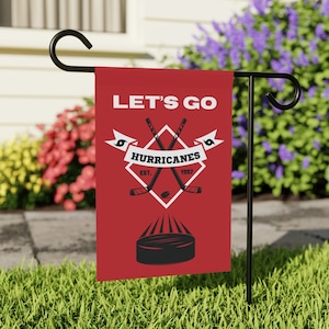 Könnte beinhalten: Eine rote Gartenflagge mit den Worten "Let's Go" und einem Hockey-Logo. Das Logo zeigt zwei gekreuzte Hockeyschläger, einen Puck und die Worte "Hurricanes Est. 1997".