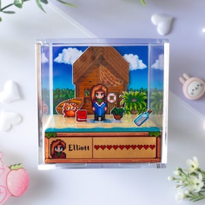 Könnte beinhalten: Eine quadratische, durchsichtige Acryl-Displaybox mit einem 3D-Pixel-Art-Diorama einer Strandszene, bestehend aus einer Holzhütte, einer Figur namens Elliott, einer Krabbe, einer Topfpflanze und einer Flaschenpost mit zehn Herzen.