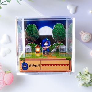 Puede incluir: Una caja de exhibición cuadrada de acrílico transparente que contiene un diorama de arte de píxeles en 3D de un personaje llamado Abigail. La escena presenta una figura de cabello azul, una linterna, una espada y árboles bajo una luna llena.