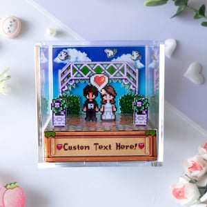 Könnte beinhalten: Ein durchsichtiger Acryl-Würfel als Schaukasten mit einer Pixel-Art-Hochzeitsszene, in der zwei Figuren unter einem Rankgitter stehen. Die Basis zeigt ein holzfarbenes Paneel mit dem Text Custom Text Here! und zwei kleinen roten Herzen.
