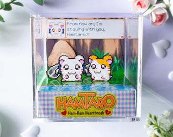 May include: A clear acrylic cube diorama featuring pixel art characters from Hamtaro: Ham-Ham Heartbreak. Inside, two hamster figures stand on a green base with a text box reading: From now on, I'm staying with you, Hamtaro. The base displays the Hamtaro logo.