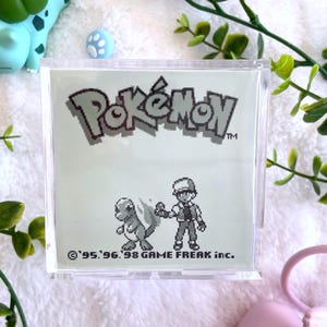 May include: A clear plastic case with a white background and a black and white image of a Pokemon character and trainer. The text "Pokemon" is printed in black and white at the top of the case. The text "'95,'96,'98 GAME FREAK inc." is printed at the bottom of the case.