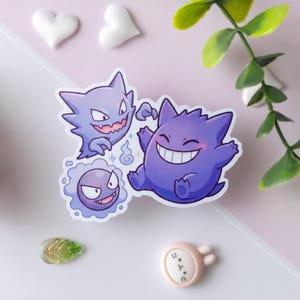 May include: A die-cut vinyl sticker featuring three purple cartoon ghost creatures. One creature is round and smiling, another is spiky with a jagged mouth, and the third is a small, circular, floating spirit. The sticker has a white border.