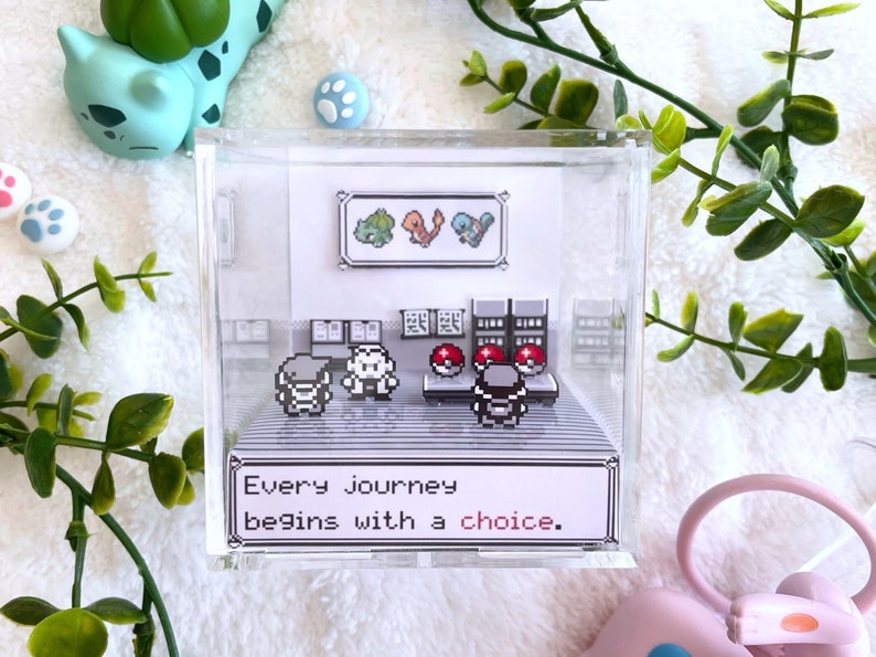 May include: A clear acrylic box with a 3D diorama of a scene from the Pokemon video game. The diorama features a trainer, a Pokemon, and a Pokeball. The box also includes a quote from the game: "Every journey begins with a choice."
