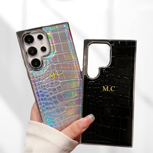 May include: Two phone cases, one silver and one black, with a crocodile skin texture. The silver case has a holographic sheen and the initials "M.C." in gold. The black case also has the initials "M.C." in gold.