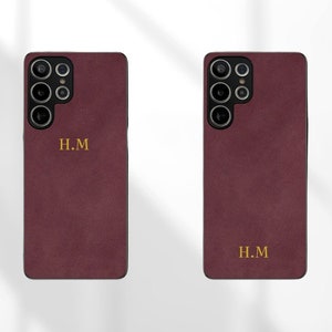 Puede incluir: Dos fundas de teléfono burdeos con recortes de cámara negros y letras doradas que dicen "H.M."