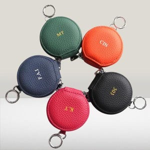 May include: Six round keyrings in a variety of colours, including green, orange, navy blue, red, and black. Each has a zip closure and a silver keyring. Initials or numbers are embossed on each in a contrasting colour.