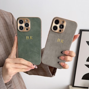 May include: Two phone cases with a suede-like texture. One is green and the other is grey. Both cases have a gold camera ring and the initials "B.K." printed in gold.
