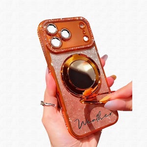 May include: Rose gold phone case with a glittery back and a ring holder. The case has a metallic frame with a diamond-like trim around the camera and edges. The word "Weather" is written on the bottom of the case.