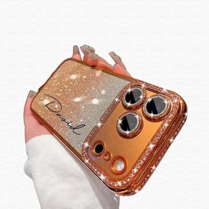 May include: A gold and silver glitter phone case with the name "David" in cursive script. The case has a gold camera frame with three lenses and is encrusted with rhinestones.