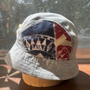 May include: A light grey bucket hat featuring a prominent patch on the front. The patch is divided into navy blue and red sections, displaying a white compass rose graphic with directional markings including NNE, NE, and ENE, and a beige letter Y.