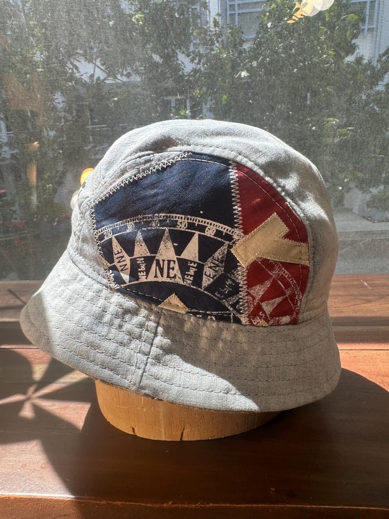 May include: A light grey bucket hat featuring a prominent patch on the front. The patch is divided into navy blue and red sections, displaying a white compass rose graphic with directional markings including NNE, NE, and ENE, and a beige letter Y.