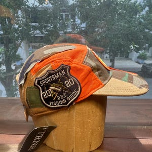 Peut inclure: Une casquette de baseball compos&eacute;e de panneaux en tissu orange vif et camouflage. Un &eacute;cusson en forme de bouclier sur le c&ocirc;t&eacute; affiche le texte SPORTSMAN 20 20 P.D.F. FISHING GUIDE avec un motif de poisson. Une &eacute;tiquette noire avec le texte TRNA est fix&eacute;e.
