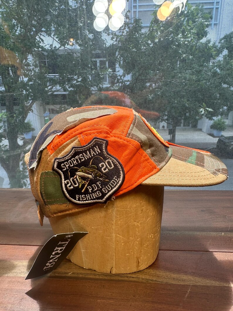 Peut inclure: Une casquette de baseball compos&eacute;e de panneaux en tissu orange vif et camouflage. Un &eacute;cusson en forme de bouclier sur le c&ocirc;t&eacute; affiche le texte SPORTSMAN 20 20 P.D.F. FISHING GUIDE avec un motif de poisson. Une &eacute;tiquette noire avec le texte TRNA est fix&eacute;e.