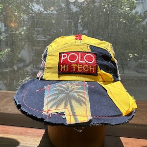 May include: A multi-paneled bucket hat featuring a yellow and navy blue color scheme with frayed edges. The front displays a rectangular patch with the text POLO HI TECH in white and yellow embroidery. A palm tree graphic is stitched onto the brim.