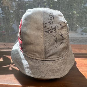 May include: A light grey bucket hat featuring a panel with a red and white plaid pattern and a section with black printed text that reads BELLE. The hat has a wide, downward-sloping brim and visible stitching along the crown and the brim edge.