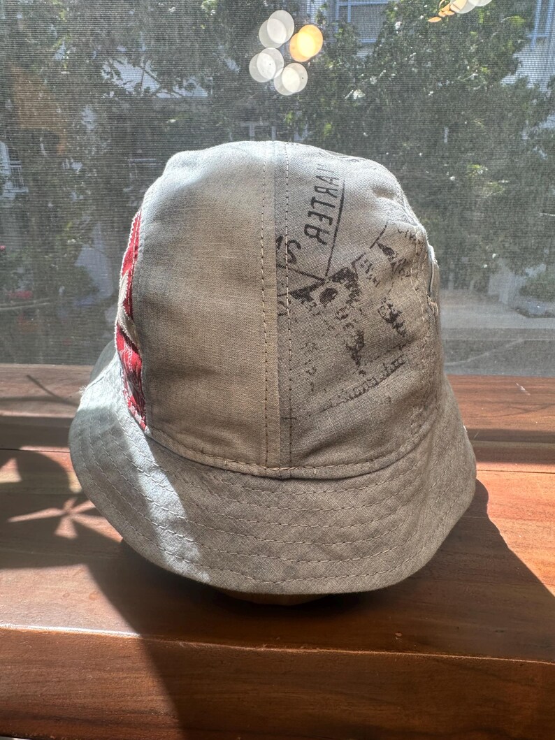 May include: A light grey bucket hat featuring a panel with a red and white plaid pattern and a section with black printed text that reads BELLE. The hat has a wide, downward-sloping brim and visible stitching along the crown and the brim edge.