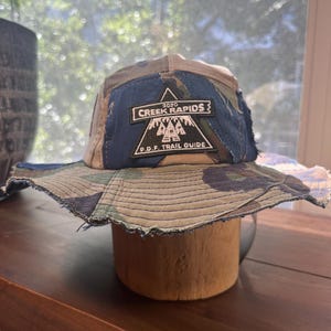 May include: A camouflage bucket hat featuring a dark blue fabric panel on the front. A black triangular patch is sewn onto the blue panel with white text that reads 2020 CREEK RAPIDS P.D.F. TRAIL GUIDE and a graphic of three pine trees.