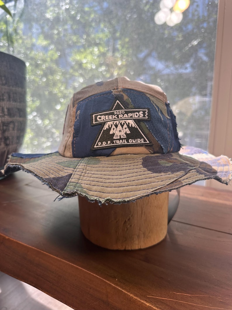 K&ouml;nnte beinhalten: Ein Camouflage-Anglerhut mit einem dunkelblauen Stoffeinsatz auf der Vorderseite. Auf dem blauen Einsatz ist ein schwarzer, dreieckiger Aufn&auml;her mit dem wei&szlig;en Text 2020 CREEK RAPIDS P.D.F. TRAIL GUIDE und einer Grafik von drei Tannen.