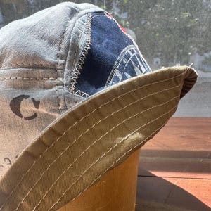 May include: A bucket hat constructed from light grey fabric with visible dark stitching. It features a dark blue triangular patch with a white graphic compass design and a wide, tan-coloured brim with multiple rows of parallel white topstitching.
