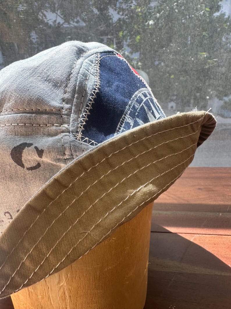 May include: A bucket hat constructed from light grey fabric with visible dark stitching. It features a dark blue triangular patch with a white graphic compass design and a wide, tan-coloured brim with multiple rows of parallel white topstitching.