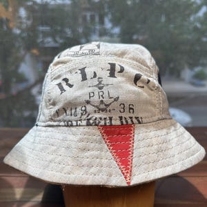 May include: A beige bucket hat featuring black printed text that reads RLPC, PRL, and 36, along with an anchor graphic. The hat includes a prominent red triangular patch on the front brim with visible white stitching and a wide, downward-sloping brim.