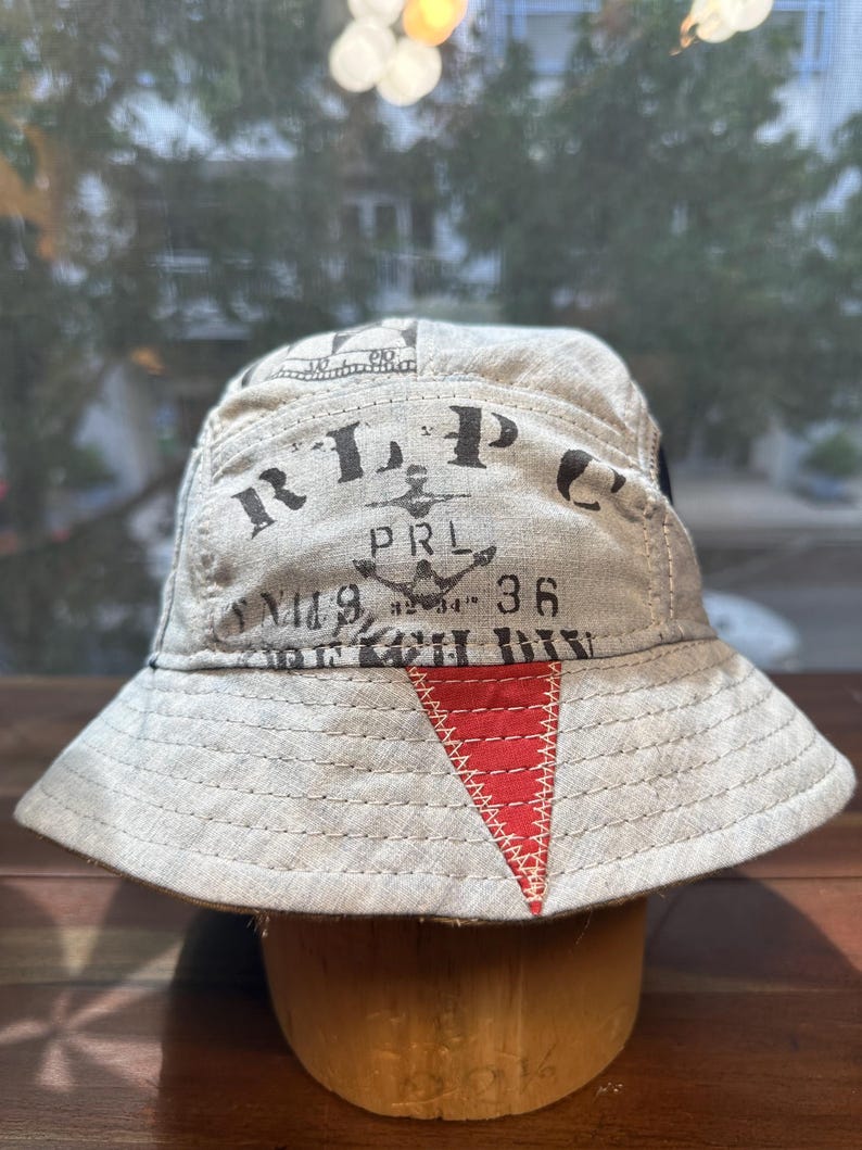 May include: A beige bucket hat featuring black printed text that reads RLPC, PRL, and 36, along with an anchor graphic. The hat includes a prominent red triangular patch on the front brim with visible white stitching and a wide, downward-sloping brim.