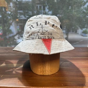 May include: A beige bucket hat featuring black printed text that reads RLP, PRL, and 36, along with an anchor graphic. The hat includes a prominent red triangular patch stitched onto the front brim and crown, with visible stitching throughout.