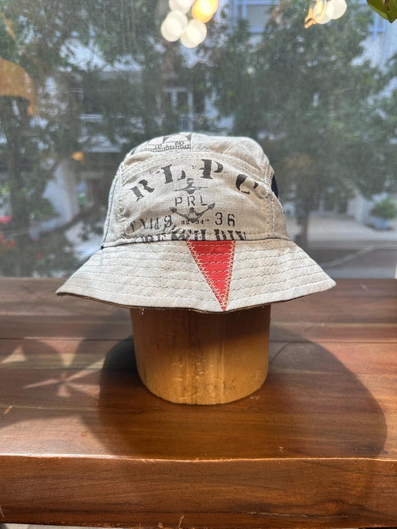 May include: A beige bucket hat featuring black printed text that reads RLP, PRL, and 36, along with an anchor graphic. The hat includes a prominent red triangular patch stitched onto the front brim and crown, with visible stitching throughout.