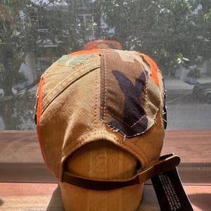 Peut inclure: Une casquette &agrave; panneaux multiples compos&eacute;e de toile beige, de tissu camouflage et d'accents orange vif. L'arri&egrave;re pr&eacute;sente une lani&egrave;re en cuir marron avec une boucle en m&eacute;tal et une &eacute;tiquette noire avec le texte blanc HANDMADE SINCE 2020.
