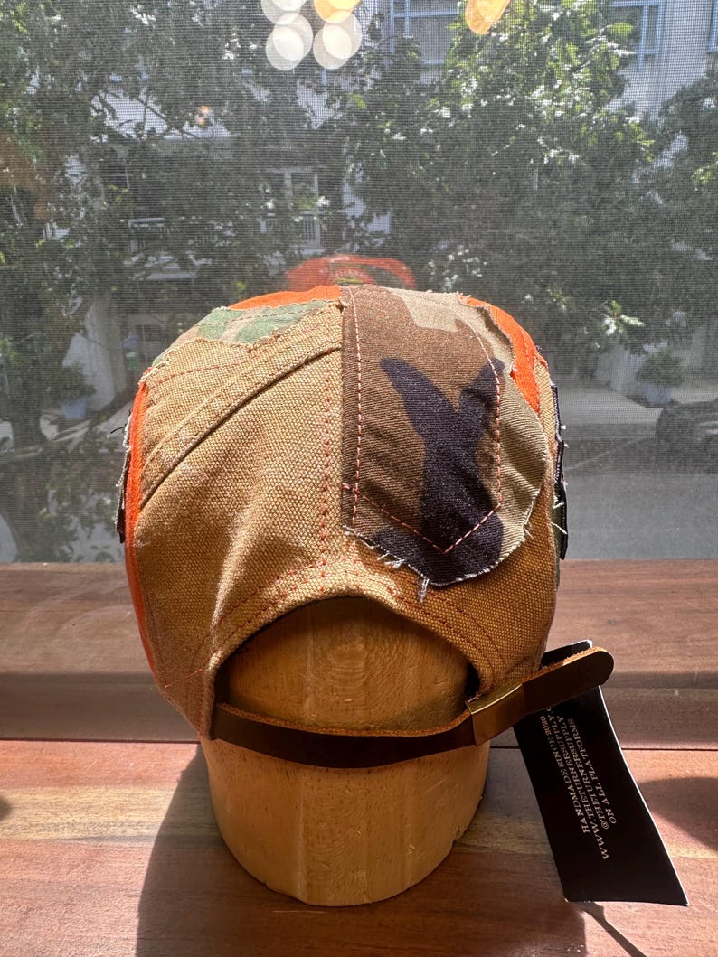 Peut inclure: Une casquette &agrave; panneaux multiples compos&eacute;e de toile beige, de tissu camouflage et d'accents orange vif. L'arri&egrave;re pr&eacute;sente une lani&egrave;re en cuir marron avec une boucle en m&eacute;tal et une &eacute;tiquette noire avec le texte blanc HANDMADE SINCE 2020.